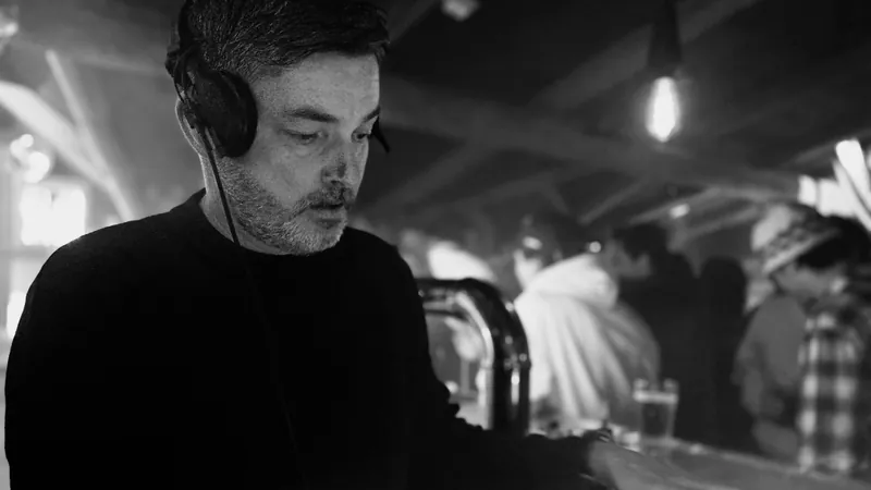 MRCIER playing a DJ set, headphones on, eyes on the decks, the crowd in the background of a smoky club